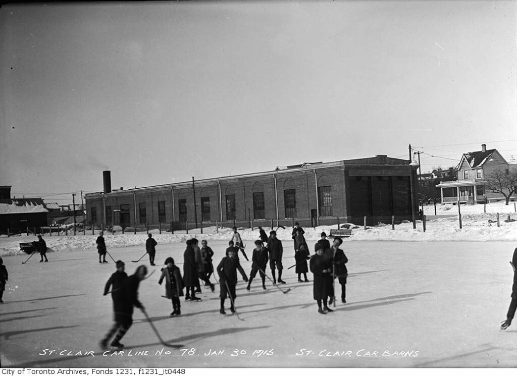archival photo of over 20 boys playing hockey forzen outdoor rink with Barns in background