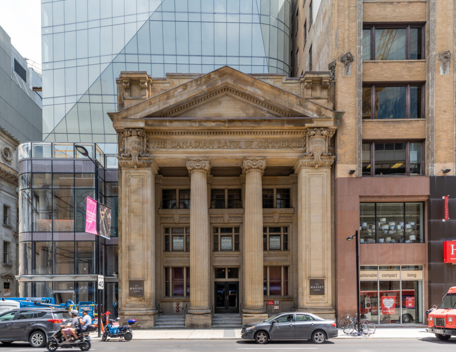 Historic Bank of Commerce building in foreground, Neoclassical style, with new glass tower behind and above. Busy Yonge Street traffic passes by. 