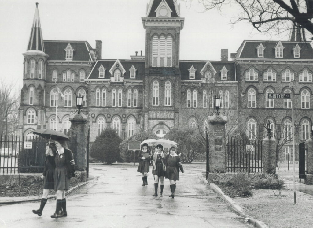 photo of teenage girls holding umbrellas walking by the entry gates of Alma College in 1983