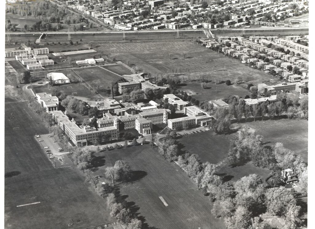 a black and white aerial photo of a cluster of buildings in the center of a grass field